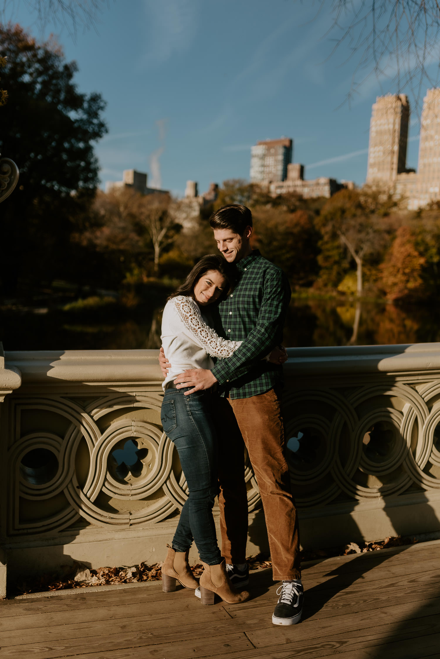 engaged couple hugs on bow bridge in front of nyc skyline at central park during their engagement photoshoot